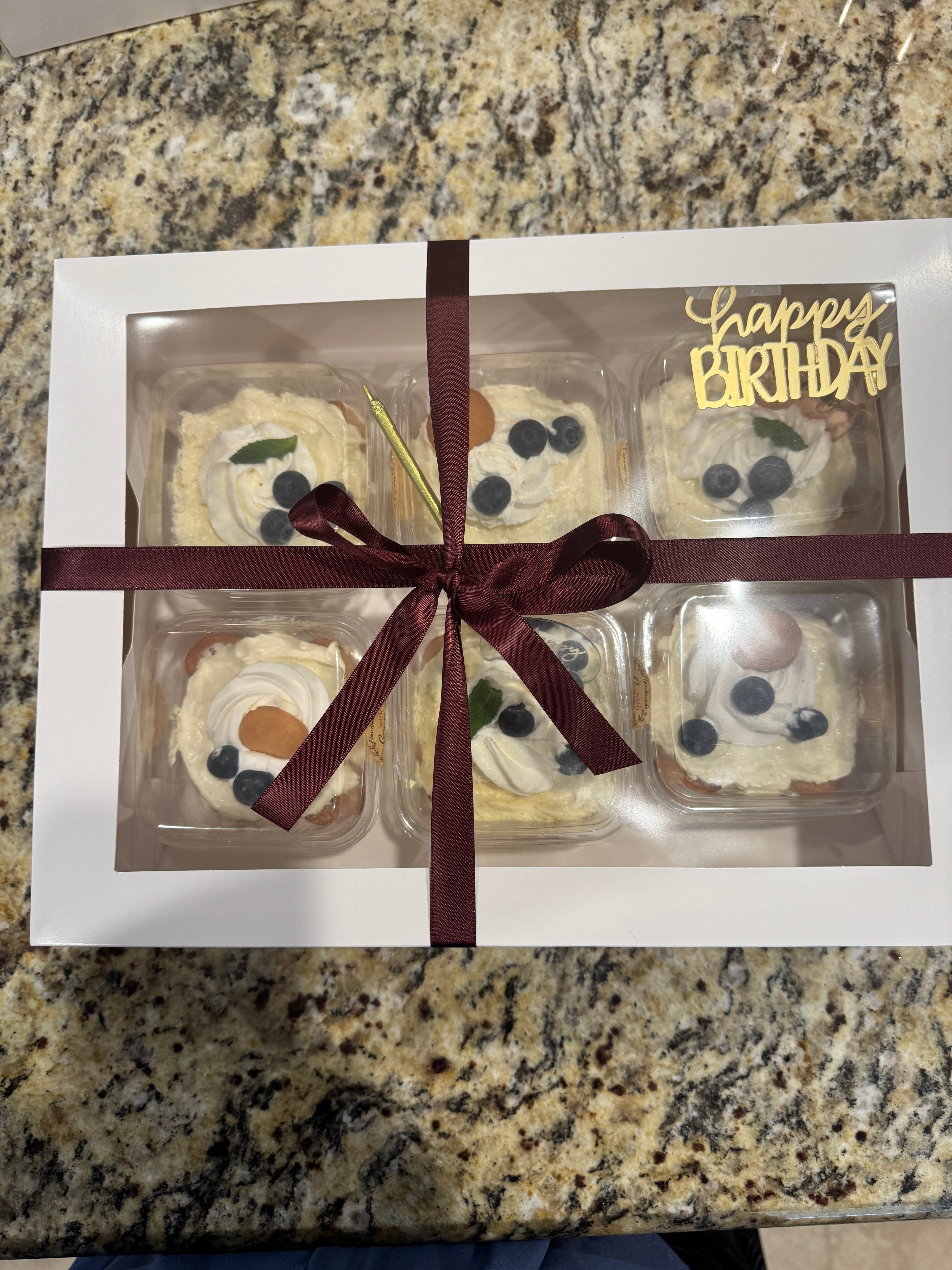A burgundy ribbon, tied into a simple bow wraps this window-bakery box. The box window is affixed with a gold-mirror script "Happy Birthday" and single gold candle. Through the window, one can see 6 clear, square, 8oz containers of banana pudding topped with whipped cream, mini nilla wafer and three blueberries.